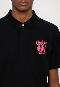 Homme portant un polo noir avec un homard rose brodé et un petit texte sur la poitrine gauche, sur un fond uni.