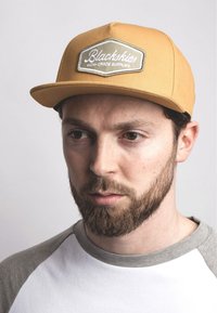 Cappello giallo con patch ricamato che riporta "Blackskies High-Grade Supplies". Maglietta grigia con scollo a costine e maniche corte.