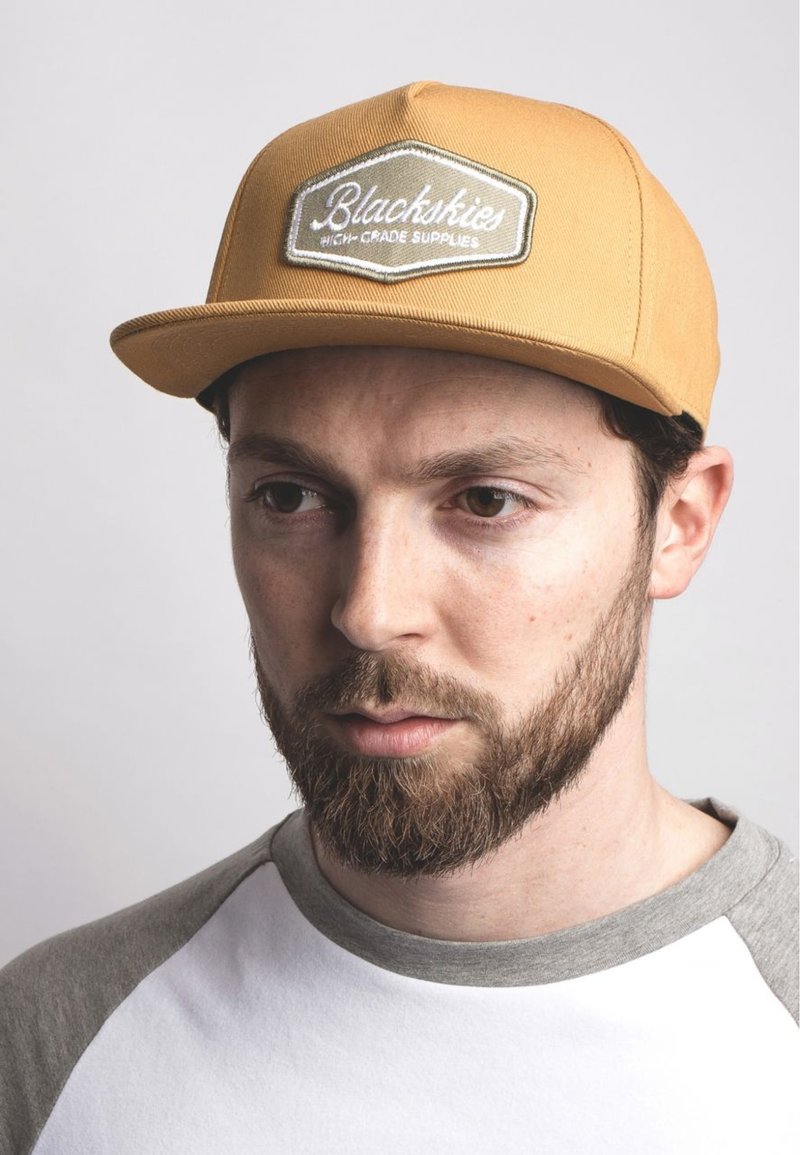 Yellow cap with embroidered patch stating "Blackskies High-Grade Supplies." Grey t-shirt with a ribbed neckline and short sleeves.