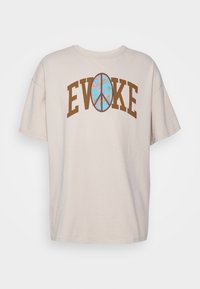 Béžové oversized tričko s krátkými rukávy, na kterém je ve středu hnědým písmem napsáno "EVOKE" a modrý znak míru s květinovými akcenty.