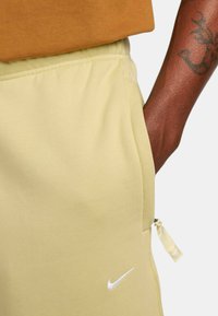 Pantaloni de jogging galbeni deschis, confecționați dintr-un material moale, cu o talie elastică, buzunar lateral cu fermoar și un logo Nike alb la margine.
