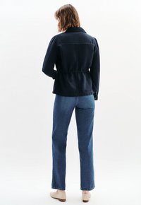 Tummansininen takki, jossa on kiristetty vyötärö, kaulus ja nappihihnat, yhdistettyinä suoriin sinisiin farkkuihin ja vaaleisiin kenkiin.