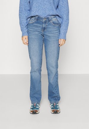 Personne portant un pull en maille bleu, un jean bleu taille moyenne et des baskets bleu sarcelle, debout sur un sol gris clair contre un mur blanc.