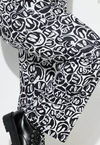 Zwart-witte, gestreepte broek met een verscheidenheid aan cartoonachtige gezichten. Het materiaal lijkt glad te zijn en heeft een reguliere pasvorm. Zwarte, grove laarzen zijn zichtbaar.