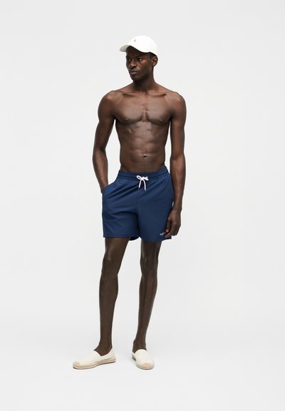 Άνδρας χωρίς πουκάμισο που φοράει ναυτικό μπλε μαγιό σορτς, άσπρο καπέλο και άσπρα παντόφλες, στέκεται με το ένα χέρι στην τσέπη σε απλό φόντο.