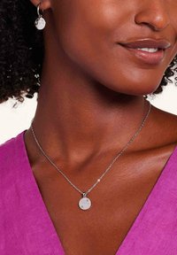 Zilveren sieraden met een ronde hanger met gegraveerde details, gecombineerd met bijpassende creolen, gedragen met een magenta top.