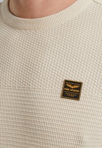 Pull en maille texturée beige avec un col côtelé et un patch logo PME Legend noir et or à l'avant.