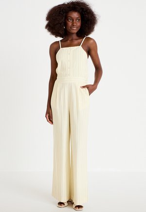 Nainen, jolla on runsaat kiharat hiukset, pukeutuneena kermanväriseen hihattomaan haalariin, jossa on pystysuorat raidat, ja sandaaleihin. Hän seisoo yhdellä kädellä taskussa.