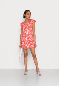 Vestido floral vermelho com mangas curtas e onduladas, com decote em V. Usado com saltos lavanda e uma bolsa a condizer, colocado contra um fundo neutro.