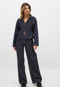 Giacca di jeans a righe blu e bianche con zip sul fronte, abbinata a pantaloni a gamba larga con un motivo coordinato e vita alta.