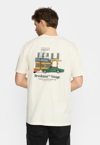 Crème katoenen T-shirt met een vintage grafisch ontwerp van gebouwen en een groene auto op de achterkant, gemarkeerd met "Revolution™ Vintage."