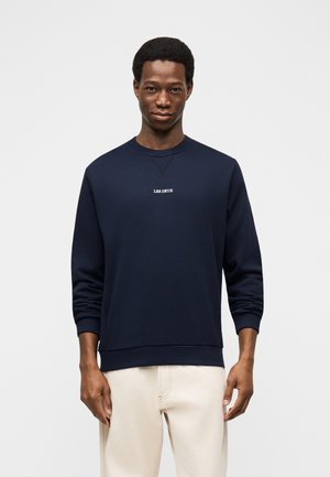 Ung mand iført en marineblå sweatshirt med teksten "LES DEUX", kombineret med lyse beige bukser, stående foran en ensfarvet hvid baggrund.