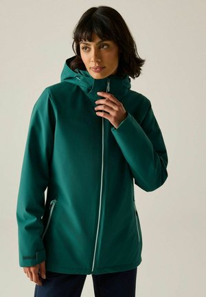 Mujer con cabello oscuro con una chaqueta con capucha color verde azulado con cremallera frontal y bolsillos laterales con cremallera, sosteniendo el cuello de la chaqueta cerca de su rostro.