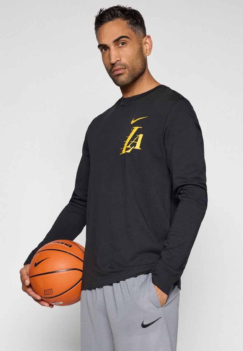 Camisa negra de manga larga con el logo amarillo de "LA" en el pecho, combinada con pantalones cortos grises. El modelo sostiene un balón de baloncesto naranja con detalles en negro.
