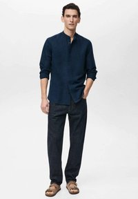 Dark blue linen button-up shirt with a stand collar, paired with dark denim pants and brown sandals. Simple, relaxed fit with rolled sleeves.