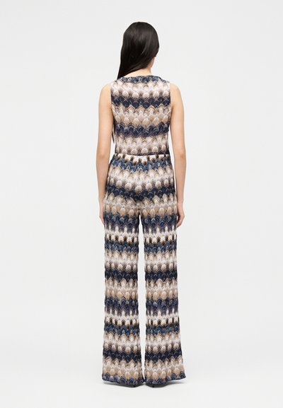 Γυναίκα με μακριά σκούρα μαλλιά που φοράει ένα αμάνικο, εμπριμέ ολόσωμο παντελόνι με φαρδιά πόδια, στέκεται με την πλάτη προς ένα απλό φόντο.