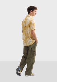 Korte mouwen gele shirt met witte tropische bladvormen, gecombineerd met olijfgroene cargo broek en sneakers met gele accenten.