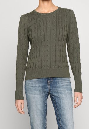 Femme portant un pull en maille torsadée vert olive à manches longues et un jean bleu clair délavé sur fond uni.