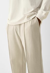 Béžové bavlněné kalhoty s řasami a bočními kapsami. Doplněné volnou bílou košilí s žebrovanými manžetami.