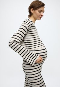 Csíkos kötött ruha bézs és fekete színekben, hosszú harang ujjakkal, testhezálló szabással és lekerekített szegéllyel, kiemelve a terhes sziluettet.