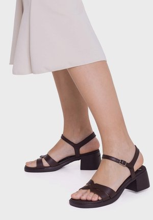 Nogi w ciemnobrązowych sandałach z odkrytymi palcami z niskimi, kwadratowymi obcasami i paskami wokół kostki, połączone z jasnobeżową spódnicą.