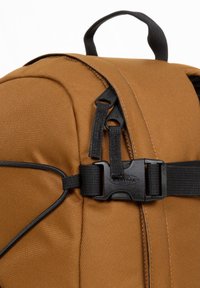 Mochila de tela marrón con correas y hebilla negras. Presenta una textura suave y costuras resistentes, tiradores de cremallera visibles y un logo.