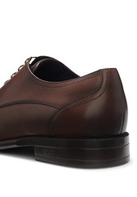 Chaussure formelle en cuir marron avec des coutures détaillées et "BOSS" embossé sur le talon de la semelle noire, vue de côté et de dos.