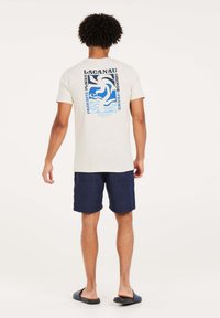 Crème t-shirt met korte mouwen met een blauw en zwart graphic ontwerp op de achterkant. G gedragen met marineblauwe shorts en zwarte sliders.