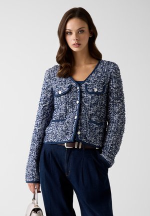 Femme portant une veste bleue texturée boutonnée avec des poches avant, un pantalon bleu foncé, tenant un sac à main blanc sur un fond uni.