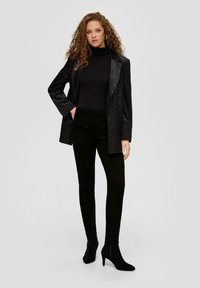 Crni teksturirani blejzer složen preko prilagođenog crnog turtleneck džempera, u kombinaciji s crnim uskim hlačama i crnim čizmama s visokom petom do gležnja.