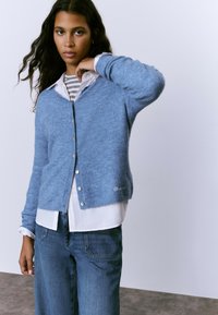 Cárdigan azul claro con una textura suave, con botones en la parte delantera, superpuesto a una camisa blanca con cuello y rayas grises.