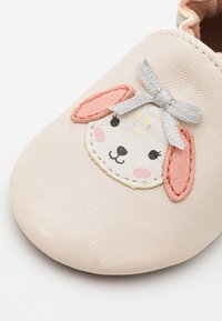 Vaaleanbeige nahkakengät, joissa on pupuvasemman muotoilu, jossa on vaaleanpunaiset korvat, mustat silmät ja harmaa nauha. Pehmeä pinta ja pyöreä muoto.