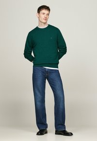 Maglione di cotone verde con collo rotondo e polsini a coste, abbinato a jeans blu scuro e scarpe nere. Fit semplice e classico.