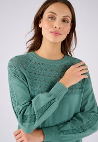 Pull en teal avec des détails en dentelle sur les épaules et les manches, doté d'un col ras du cou et de poignets côtelés. Matière douce et légère.