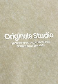 Tela de algodón de color beige claro con una textura suave que presenta texto impreso en blanco: "Originals Studio", "Presentado por JACK&JONES" y "Diseñado en Copenhague".