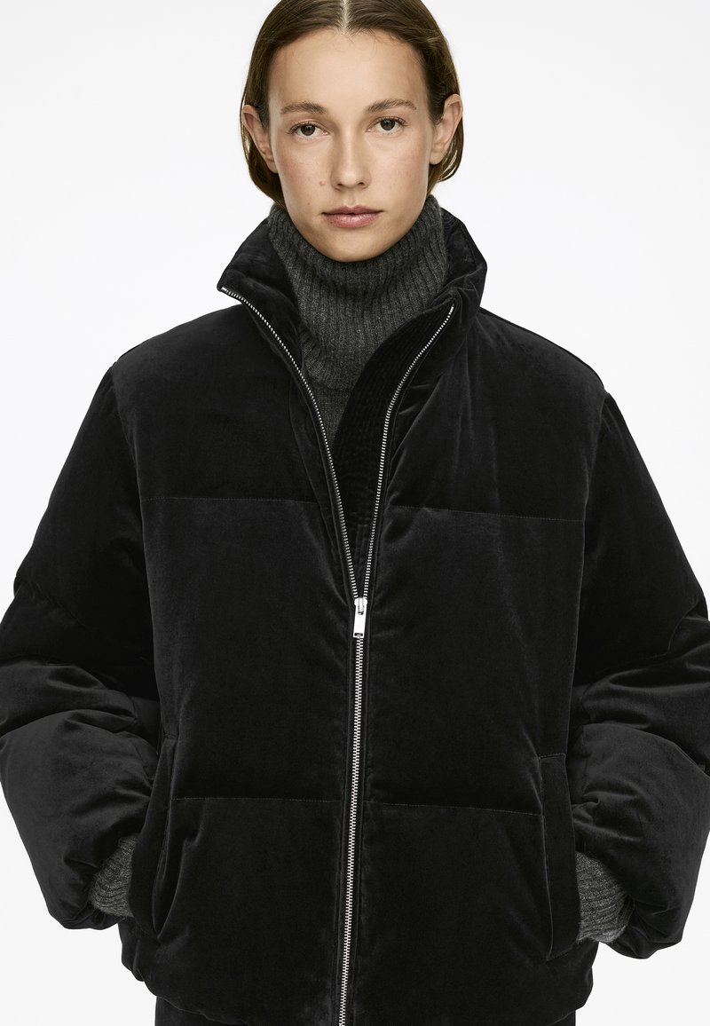 Sort fløjls-pufferjakke med høj krave, lynlås-lukning, oversized ærmer og tekstureret grå turtleneck. Glat, blød finish med subtil glans.