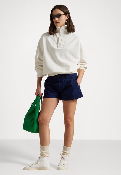 Weißer Sweater mit hohem Kragen und Knopfdetails, kombiniert mit marineblauen Shorts. Die Accessoires umfassen eine grüne Tasche und cremefarbene Sneaker in High-Top-Form.
