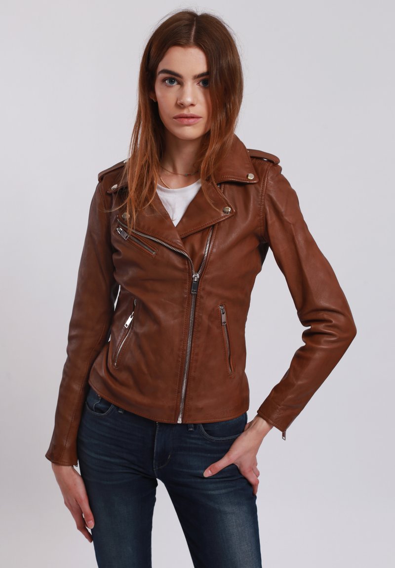 Braune Lederbikerjacke mit silbernen Reißverschlüssen, asymmetrischer Vorderseite und Druckknopfkragen-Details, kombiniert mit einem weißen Oberteil und dunklen Jeans.