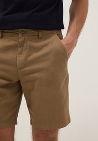 Hombre vestido con pantalones cortos marrones hasta la rodilla con la mano en el bolsillo, combinado con una camisa negra, de pie contra un fondo liso.