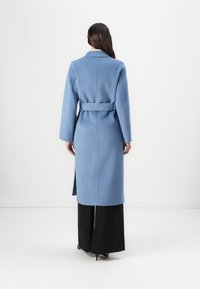 Długi niebieski płaszcz z miękkiej tkaniny z taliowaniem, bocznymi rozcięciami i klasycznym kołnierzem, zestawiony z czarnymi spodniami o szerokich nogawkach.