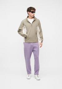 Νεαρός άντρας με μαύρα γυαλιά ηλίου, μπεζ μπουφάν με φερμουάρ, λευκό πουκάμισο, λιλά παντελόνι φόρμας και λευκά αθλητικά παπούτσια, στέκεται με το ένα χέρι στην τσέπη.