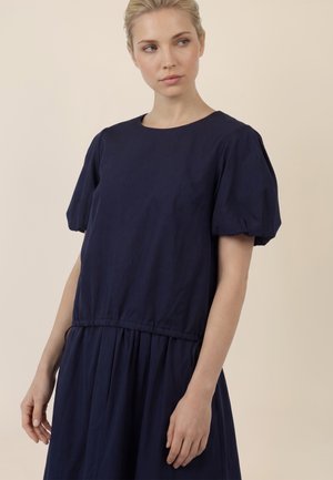 Frau mit blondem Haar, die ein marineblaues Kleid mit angesetzten Puffärmeln und geraffter Taille trägt, schaut leicht zur Seite.