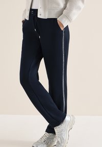 Personne portant un jogger bleu marine avec des rayures latérales blanches à motifs, des baskets blanches et une veste de couleur claire, les mains dans les poches.
