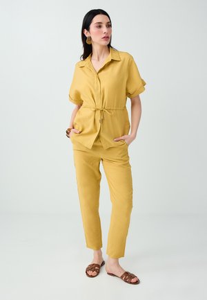 Vrouw staat met de handen in de zakken, draagt een geel kort mouwen overhemd met ceintuur en bijpassende broek, bruine sandalen en opvallende oorbellen.