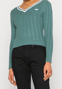 Teal knitted sweater with a V-neck, ribbed cuffs, and hem. Features white accents and a small logo on the chest. Paired with black pants.