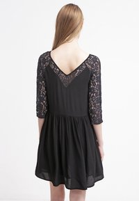 Vestido negro con mangas de encaje de tres cuartos y diseño de espalda en V. La falda está fruncida para obtener una forma fluida y una textura suave.