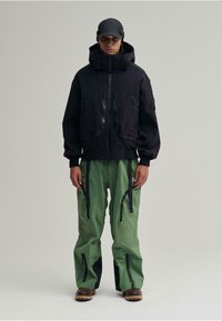 Schwarze isolierte Jacke mit hohem Kragen und Reißverschlusstaschen, kombiniert mit grünen wasserdichten Hosen mit schwarzen Akzenten und mehreren Reißverschlüssen.