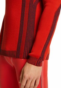 Pull en maille rouge avec des rayures horizontales foncées, doté de poignets et d'un ourlet côtelés. Associé à des leggings rouges ajustés avec des accents texturés.