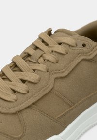 Nærbilde av en lysebrun semsket sneakers med tykke matchende snører og hvit gummisåle på ensfarget bakgrunn.