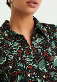 Närbild av en person som bär en svart skjorta med knappar som har gröna och rostfärgade blommönster, och guldörhängen synliga i ena örat.